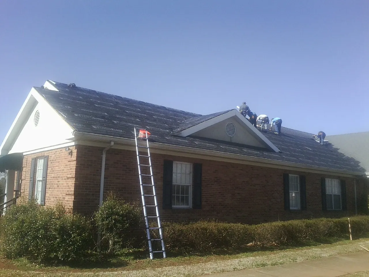 Crew tearing shingles off a brick ranch roof with a ladder set — Emergency Roof Repair work by our crew in Hickory Grove