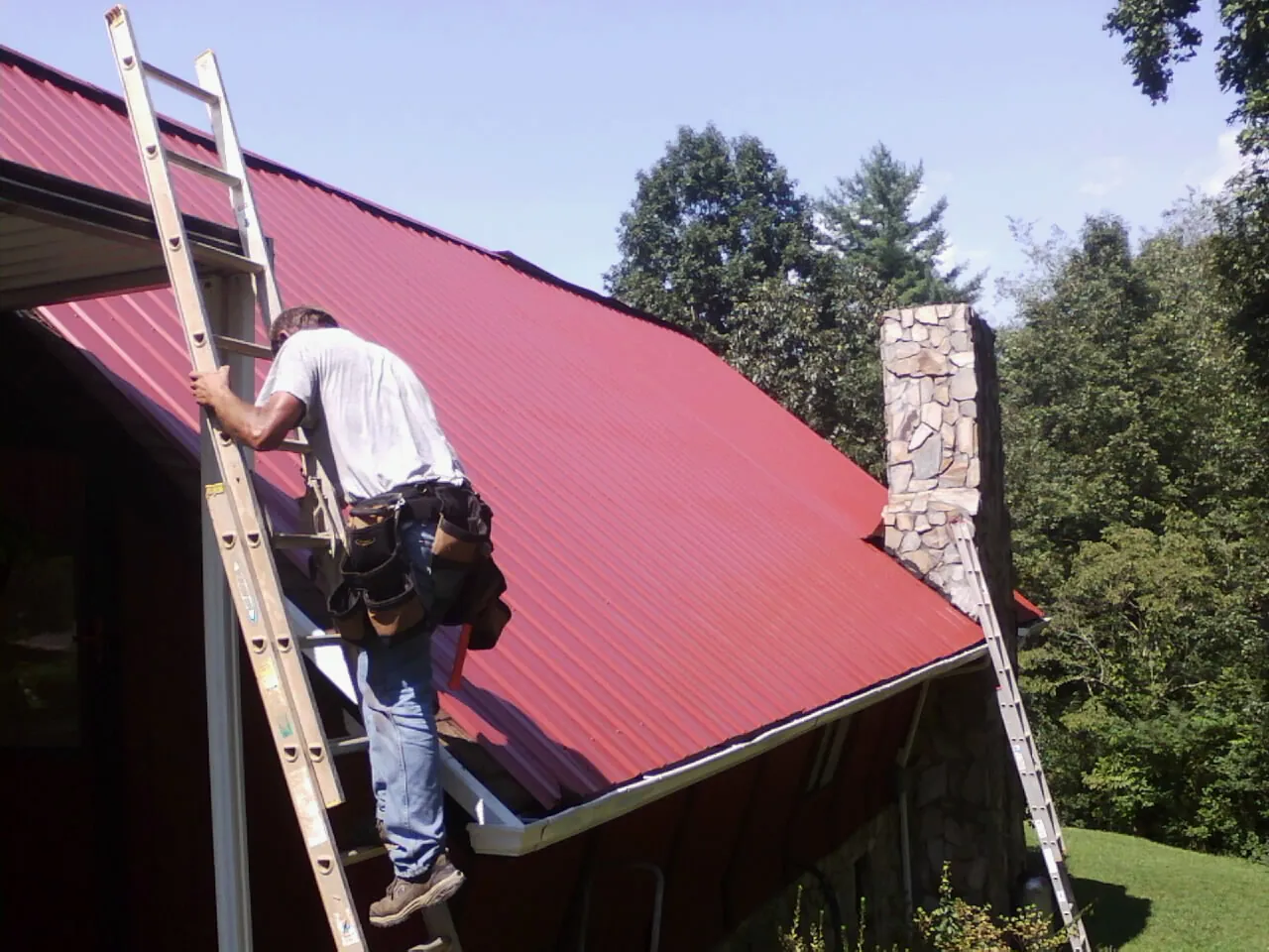 Roofer on a ladder beside a red metal roof and stone chimney — Soffit & Fascia Repair by Roofing Brothers in Hickory Grove